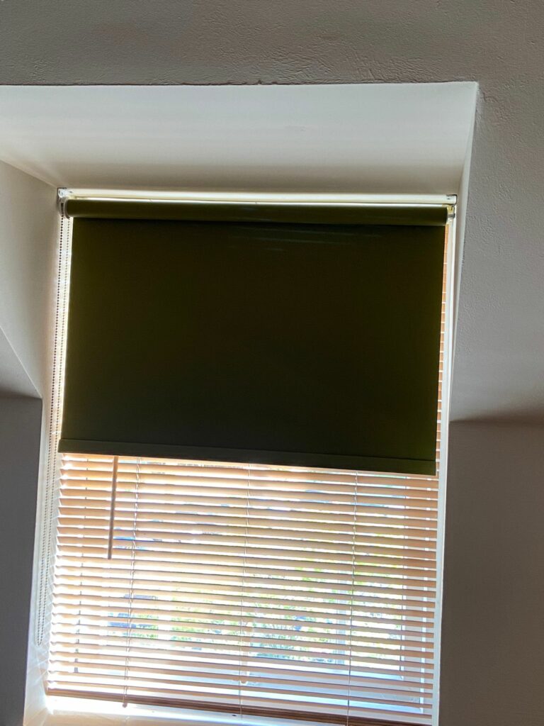 Dark green roller blind installed in a Wellingborough home, adding warmth, privacy and a modern finish while complementing the natural wood Venetian blinds below.